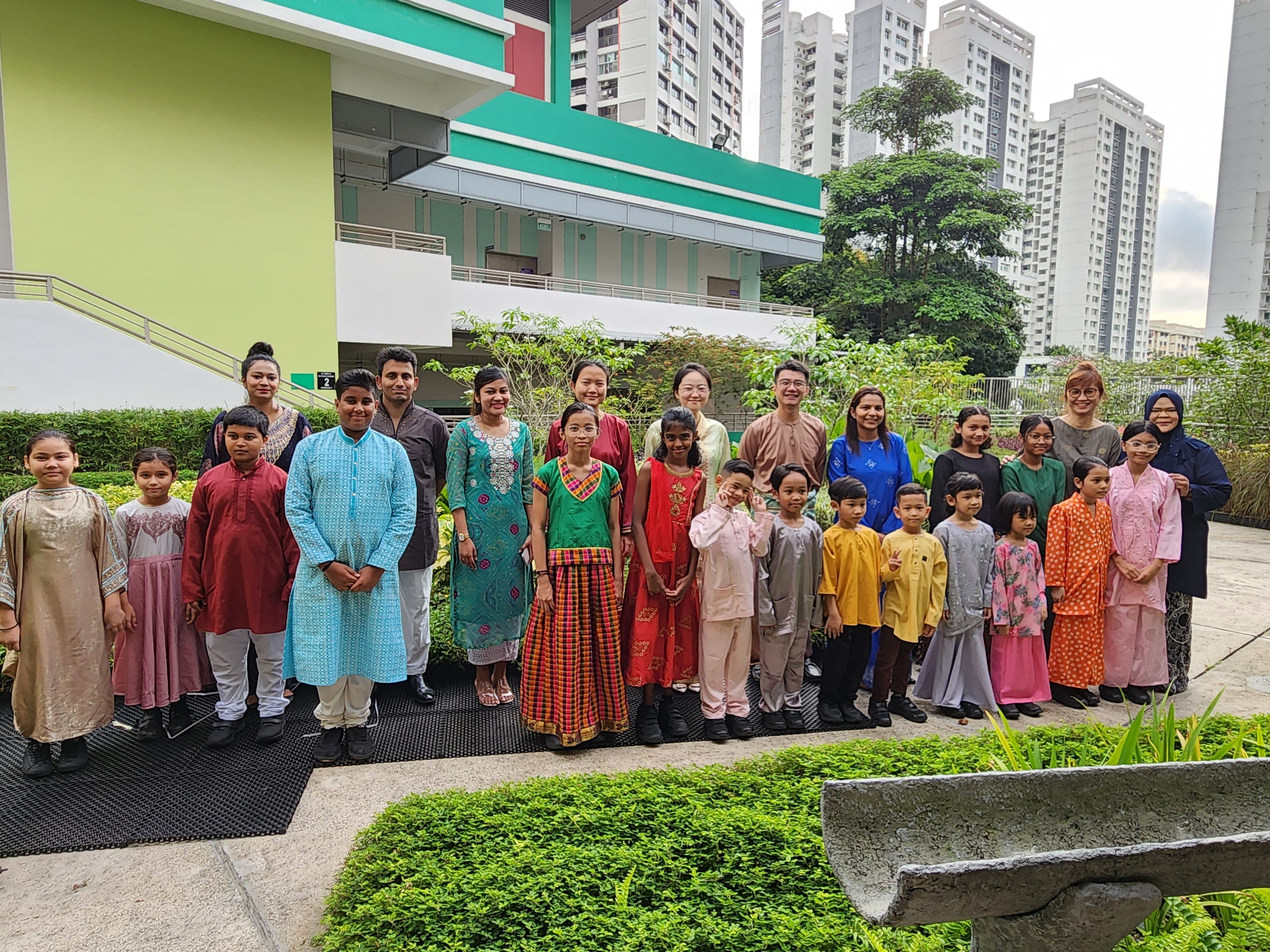 A large group of adults and children in colorful traditional South and Southeast Asian attire pose outdoors near a building.